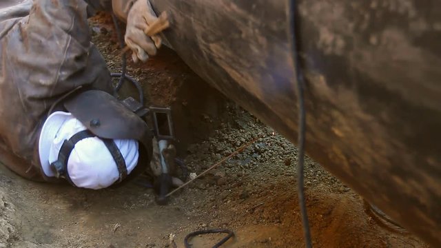 Welder is in trench working hard, arc welding pipeline.