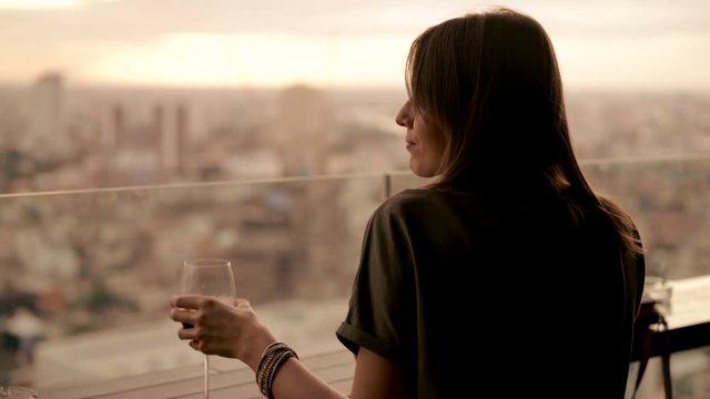Happy woman drinking red wine while sitting in rooft bar in the city, 4K
