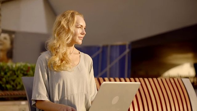 woman is writer freelancer is sitting on a street, watching a sunset , holding laptop on knees