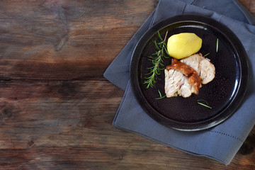 Slices of roast pork with rosemary and potato on a plate, dark rustic wooden background with generous copy space, top view from above