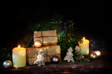 gift boxes in kraft paper and two burning candles decorated with christmas balls, fir branches and small wooden trees on a dark rustic background with copy space
