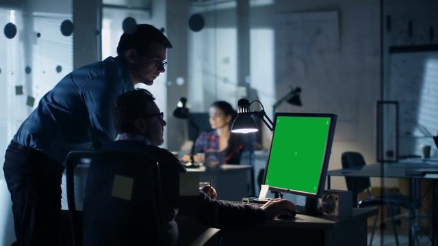 Late at Night Two Industrial Engineers Have Discussion, Personal Computer Shows Isolated Green Screen Mock-up Display. Office Looks Modern with Lots of Blueprints on the Walls.