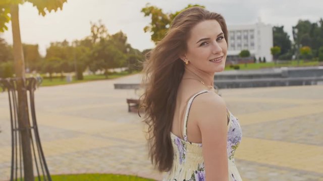 A charming sensual young girl in a summer dress is walking along the park in the center of the city. Her long hair flies in the wind. Enjoying a summer's day. The youth. Beauty.