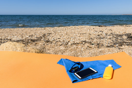 Sunglasses lying next to a smartphone on a microfiber towel next to the sunscreen on the travel rug on the beach with sea views