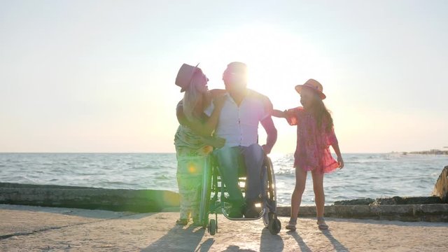 Joyful Family with paralysis man in wheelchair hugging each other lay their hands on the belly of a pregnant mother On background sea