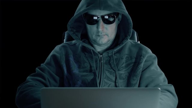 A man in hood and glasses sitting at table and coding on laptop. Dark night. Hacker in glasses working at computer. Coding man at concrete background. Alpha channel chroma key transparent background.