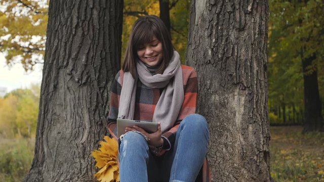 Wonderful autumnal view. Charming young happy smiling woman sitting on the grass in a park, use tablet