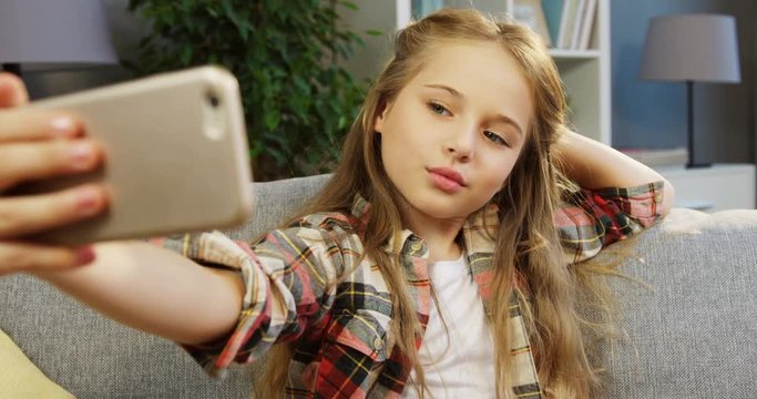 A little blonde cute girl making funny selfies on the smart phone and posing while sitting on the couch. Indoors