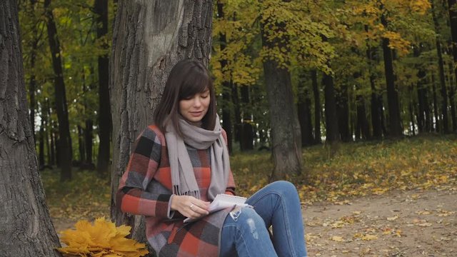 take a leaf from the notebook. Charming young happy smiling woman sitting on the grass in a park, take notes in notebook.