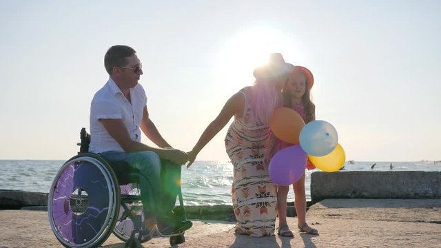 happy daughter sitting on handicapped dad in wheelchair with balloons in hand, parents with child hugging in backlight