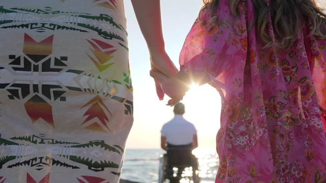 mother and daughter keep hands on background father sitting in wheelchair near water