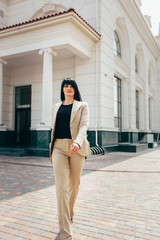 a thin girl with a dark long straight hair in a beige strict suit are standing in the full growth near the building with an amazing architecture