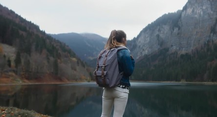 DOLLY IN Beautiful Caucasian female hiker with backpack standing near beautiful lake in Alps, rainy weather, morning shot. 4K UHD 60 FPS SLO MO