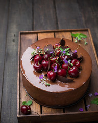 A piece of chocolate cherry mousse cake is decorated with lilac flowers and fresh cherries on a wooden box on a wooden dark background.  Dark food photo