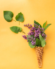 Ice cream cone with lilac branches on a bright yellow background. Fresh flowers. beautiful bouquet of flowers.  Copy Space. View blank space for text. Place for text. Food photo. Flat lay, top view