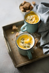 Pumpkin soup with coconut milk, pumpkin seeds and spices in vintage blue mugs on a gray concrete background. Atmospheric dark food photo. 