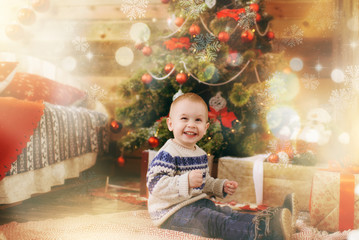 Happy little baby boy dressed in sweater decorating Christmas tree with toys in wooden room at home. Child good mood. New Year. Lifestyle, family and holiday 2018 concept. Magical highlight effect