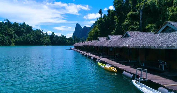 Aerial: Several bungalows standing on a lake with thatched roofs. Kayaks are parked to the pier. Cheow Lan lake. Khao Sok. Thailand.