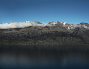 Paisaje de montañas con picos nevados y nubes. Las montañas se reflejan en un lago. Escena diurna, cielo azul y despejado.