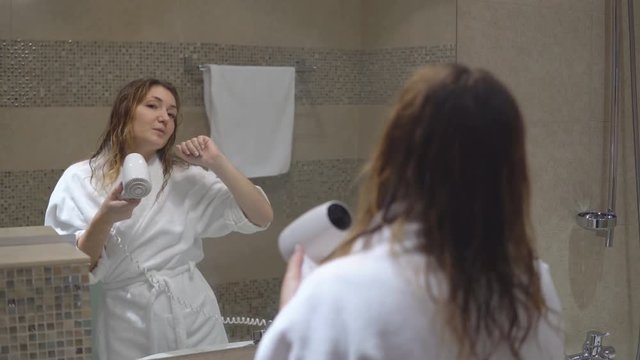 Funny charming girl in white robe took bath and sings in front of mirror with hairdryer