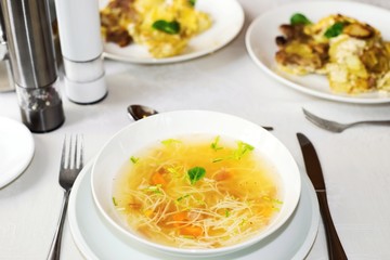 Lunch or dinner table with white plate with beef broth with noodle and two plates with baked potato, cutlery and caster.