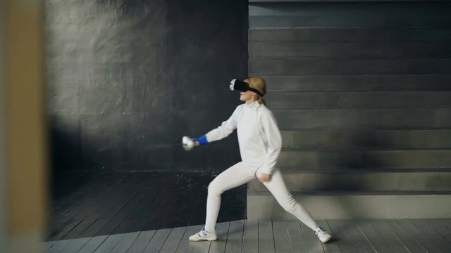Concentrated fencer woman using virtual reality headset for play fencing training simulator game indoors