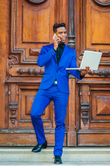 African American Businessman works in New York. Wearing blue suit, black shirt, bow tie, leather shoes, black man walks down stairs of vintage office doorway, works on laptop computer, talks on phone