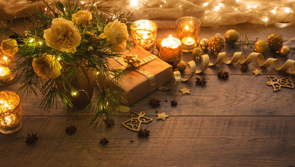 Christmas decoration. Gift, candles, lights, golden balls on a wooden rustic table. Xmas composition of pine branches and English roses in a vase. Golden and brownish aesthetics.