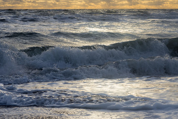 Small waves in a troubled sea roll on the shore with foam and spray under the cloudy sky and the rays of the sun