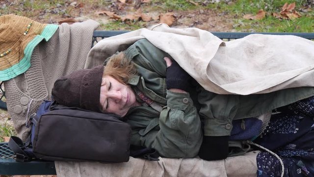 Cold homeless woman wrapped in old blanket asleep on park bench