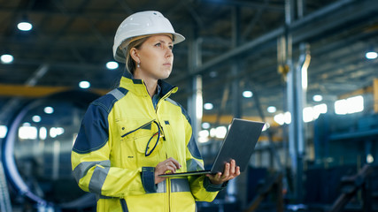 Female Industrial Engineer in the Hard Hat Uses Laptop Computer while Standing in the Heavy...