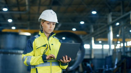 Female Industrial Engineer in the Hard Hat Uses Laptop Computer while Standing in the Heavy Industry Manufacturing Factory. In the Background Various Metalwork Project Parts Lying