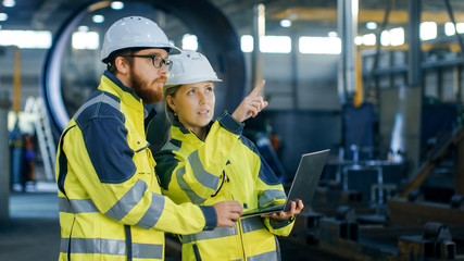 Portrait of Male and Female Industrial Engineers in Hard Hats Discuss New Project while Using Laptop. They Wear Safety Jackets.They Work at the Heavy Industry Manufacturing Factory.