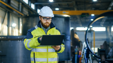 Industrial Engineer in Hard Hat Wearing Safety Jacket Uses Laptop. He Works in the Heavy Industry Manufacturing Factory with Various Metalworking Processes are in Progress.