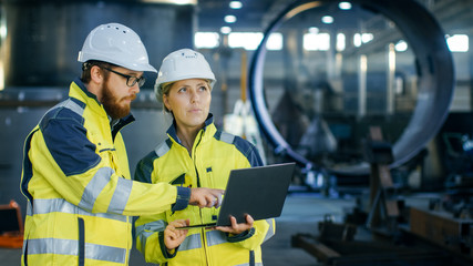 Male and Female Industrial Engineers in Hard Hats Discuss New Project while Using Laptop. They Make...
