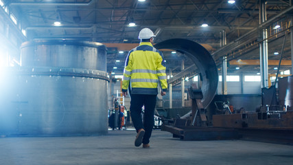 Industrial Engineer in Hard Hat Wearing Safety Jacket Walks Through Heavy Industry Manufacturing Factory with Various Metalworking Processes.