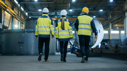 Following Shot of Three Engineers Walking Through Heavy Industry Manufacturing Factory. In the...