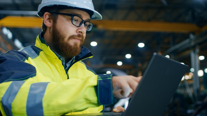 Industrial Engineer in Hard Hat Wearing Safety Jacket Uses Touchscreen Laptop. He Works at the Heavy Industry Manufacturing Factory.