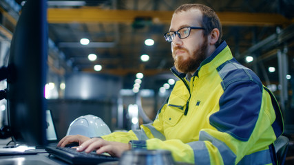 Industrial Engineer Works at Workspace on a Personal Computer.  He Wears  Safety Jacket and Works in the Main Workshop of the Heavy Industry Manufacturing Factory.