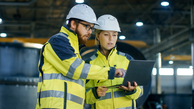 Male and Female Industrial Engineers in Hard Hats Discuss New Project while Using Laptop. They Make Showing Gestures.They Work at the Heavy Industry Manufacturing Factory.