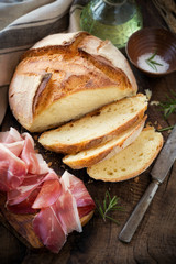 Freshly baked homemade bread on dark rustic wooden table with sliced prosciutto ham, salt and olive oil at the background