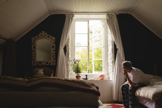 Man looking through window while sitting on bed at home