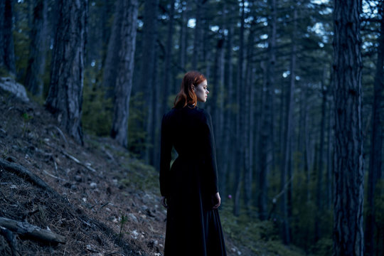 Young beautiful woman in a long black dress is standing in a dark forest