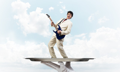 Businessman on metal tray playing electric guitar against blue sky background