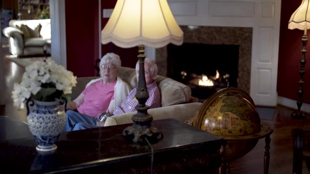 Elderly couple sitting on a couch smiling with a fire in the fireplace.