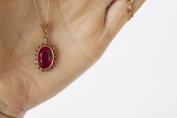 The hand holds a gold chain and a gold pendant with a ruby