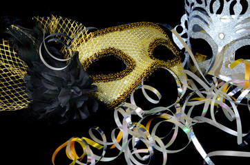 New Year's Eve themed image of gold and silver carnival masks decorated with black flower and feathers with curly ribbons on black background.