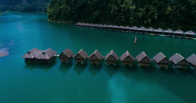 Aerial: The hostel on the lake with thatched roofs houses on the water. Kayaks are parked to the pier. Cheow Lan lake. Khao Sok. Thailand.