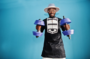 Portrait of stylish black african american man at hat and glasses with dumbbells in his  hands against blue background.