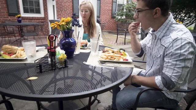Mature mother and hipster teen son sit at an outdoor table and eat a lunch of meatball subs with fries.